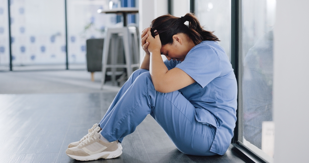 Caregiver burnout in Brooklyn shown through an exhausted woman in scrubs sitting on the floor